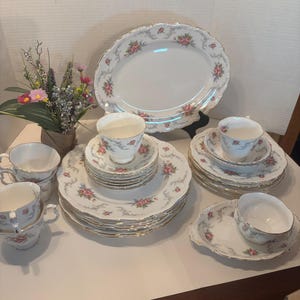 May include: A vintage porcelain tea set with a floral rose design and gold edging. The set includes teacups, saucers, plates, and a serving platter. The dishes are white with pink roses and a scalloped edge.