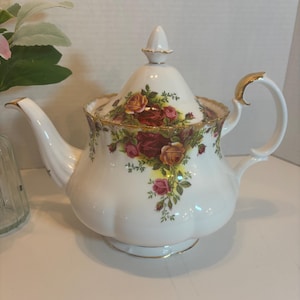 May include: White porcelain teapot with a floral rose pattern in shades of red, yellow, and pink. The teapot has a gold-trimmed handle and spout, and a matching lid with a finial. A classic tea set item.