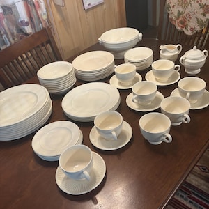 May include: A large set of cream-coloured ceramic dinnerware featuring a subtle ribbed texture along the edges. The collection includes multiple stacks of dinner plates, salad plates, and bowls, along with several teacups with matching saucers, a lidded sugar bowl, a creamer, and a large lidded tureen.