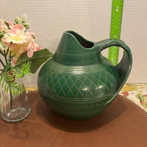 以下が含まれることがあります： ダイヤモンド模様と取っ手がついた緑色の陶器のピッチャー。ピッチャーは茶色の革の表面の上に置かれています。