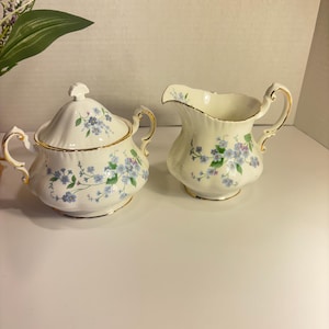 May include: A cream-coloured sugar bowl with a lid and a creamer. Both items feature blue floral patterns and gold trim. The sugar bowl has two handles and a lid knob. The creamer has a handle and a spout, typical of a tea set.