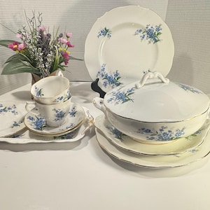 May include: A white porcelain dinnerware set featuring delicate blue floral patterns and gold trim. The collection includes a lidded tureen, dinner plates, saucers, and teacups, all decorated with matching blue blossoms and green foliage accents.