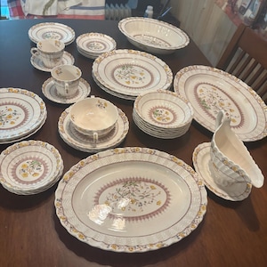 May include: A collection of white ceramic dinnerware with a floral design in yellow, purple, and green. The set includes plates, bowls, cups, saucers, and serving platters. The edges have a scalloped design.