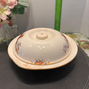 Peut inclure: Un plat de service en céramique blanche avec un couvercle. Le plat est décoré d'un motif floral dans des tons de rouge, bleu et jaune. Le couvercle a un petit bouton sur le dessus.