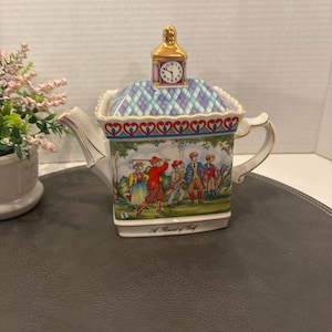 May include: A white porcelain teapot with a clock tower lid and a scene of people playing golf. The teapot has a blue and white checked pattern and a red heart border. The text "A Round of Golf" is printed on the bottom.