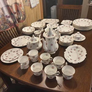 May include: A large collection of white porcelain dinnerware featuring a delicate pink floral pattern and gold trim. The set includes a tall teapot, a smaller teapot, multiple teacups, saucers, various plates, a lidded sugar bowl, and serving dishes.
