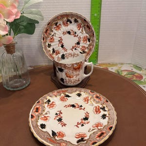 Peut inclure: Un ensemble de tasse et soucoupe à thé vintage avec un motif floral dans les tons rouge, noir et or. La tasse et la soucoupe ont un fond blanc avec un bord festonné.