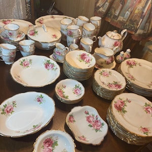May include: A large collection of white porcelain dinnerware featuring a pink rose floral pattern and gold trim. The set includes various plates, bowls, teacups, saucers, a teapot, a lidded tureen, a creamer, a sugar bowl, and salt and pepper shakers.
