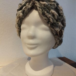 May include: Grey and white knitted headband with a black button on top. The headband is on a white mannequin head.