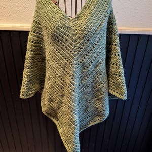 May include: A light green, handmade crochet poncho with a V-neck and open stitch detailing. The poncho is displayed on a decorative metal stand, showcasing its asymmetrical hem and wide sleeves. The garment's texture and color suggest a cozy, handcrafted aesthetic.