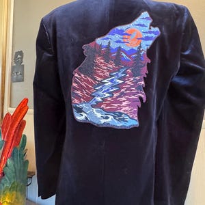 May include: A dark blue velvet blazer with a large embroidered patch on the back. The patch depicts a wolf's head with a mountain scene inside. The scene includes a river, trees, and a full moon.