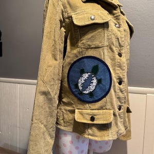 May include: A tan corduroy jacket with a blue and green patch featuring a lightning bolt and a turtle. The patch is on the left side of the jacket.