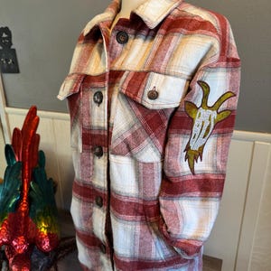 May include: A red and white plaid flannel shirt with a brown goat graphic on the left sleeve.