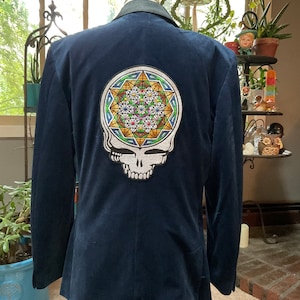 Puede incluir: Una chaqueta de terciopelo azul marino con un gran parche bordado en la espalda. El parche presenta una calavera blanca con un diseño geométrico colorido en su interior.