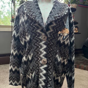 May include: A black and white chevron patterned cardigan with a brown button closure and a large embroidered animal paw print on the right side.