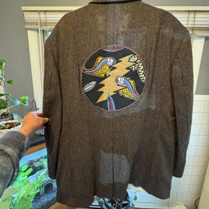 May include: A brown tweed blazer with a circular patch on the back. The patch features a lightning bolt design with two fish swimming around it. The fish are yellow, blue, and red. The patch is surrounded by a brown stitched border.