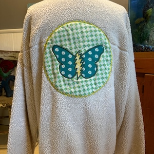 May include: A white fleece jacket with a green and white butterfly patch on the back. The butterfly has a yellow lightning bolt in the center.