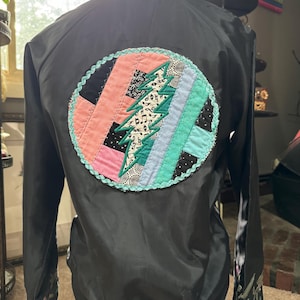 May include: Black bomber jacket with a large, colorful, patchwork design on the back. The design features a green tree with musical notes and a pink, blue, and green color scheme.