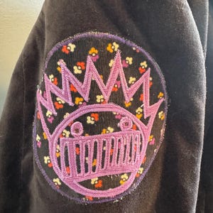 May include: A black velvet jacket with a purple and white embroidered patch on the sleeve. The patch features a crown, a face, and a musical instrument. The background of the patch is a floral pattern.