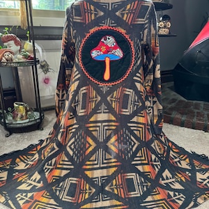 May include: A long black cardigan with a geometric pattern in orange, brown, and beige. The cardigan has a large embroidered patch on the back featuring a mushroom with a red and white top, a blue stem, and a brown base. The mushroom is surrounded by a black circle with an orange border.