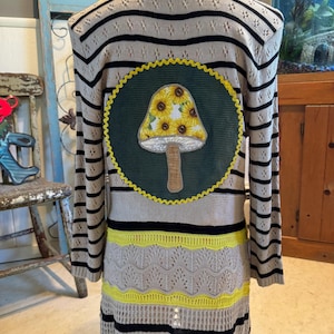 May include: A long-sleeved, striped cardigan with a large, green circle on the back. The circle features a brown mushroom with a yellow top decorated with sunflowers.