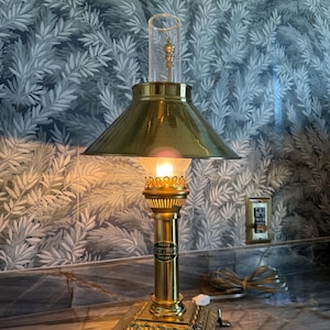 May include: A brass table lamp with a conical shade and a clear glass chimney. The lamp has a square base with decorative feet and a label that reads "PARIS". The lamp is lit, casting a warm glow.
