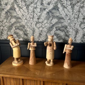 May include: Four wooden nutcracker figurines in different poses. The figures are mainly light brown and beige, with details such as hats and aprons. They are displayed on a wooden cabinet against a leaf-patterned wallpaper.