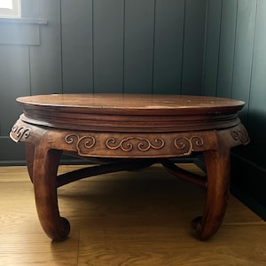 May include: A round, wooden antique table with a dark brown finish. The table features carved scrollwork along the apron and curved legs. The table is approximately 61 cm in diameter and 30 cm tall.