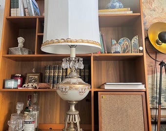 May include: A vintage table lamp with a white fabric lampshade trimmed with gold braid. The lamp has a white glass base with gold accents and crystal drops hanging from the base.