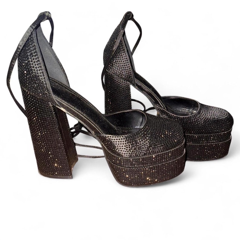K&ouml;nnte beinhalten: Ein Paar schwarze Plateau-High Heels mit funkelnden Verzierungen. Die Schuhe haben einen Blockabsatz und Kn&ouml;chelriemen. Die Plateauschuhe haben eine runde Spitze und eine hohe Plateausohle. Das Design ist glamour&ouml;s.