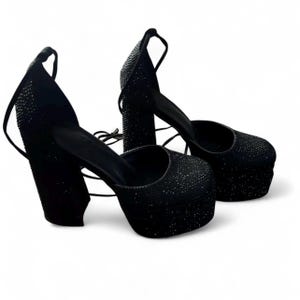 K&ouml;nnte beinhalten: Ein Paar schwarze Plateau-High Heels mit Kn&ouml;chelriemen. Die Schuhe haben eine dicke Plateausohle und einen klobigen Absatz, die beide mit kleinen, glitzernden Verzierungen bedeckt sind. Sie haben eine runde Spitze und einen Kn&ouml;chelriemen.