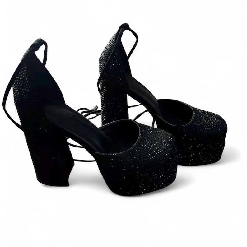 K&ouml;nnte beinhalten: Ein Paar schwarze Plateau-High Heels mit Kn&ouml;chelriemen. Die Schuhe haben eine dicke Plateausohle und einen klobigen Absatz, die beide mit kleinen, glitzernden Verzierungen bedeckt sind. Sie haben eine runde Spitze und einen Kn&ouml;chelriemen.