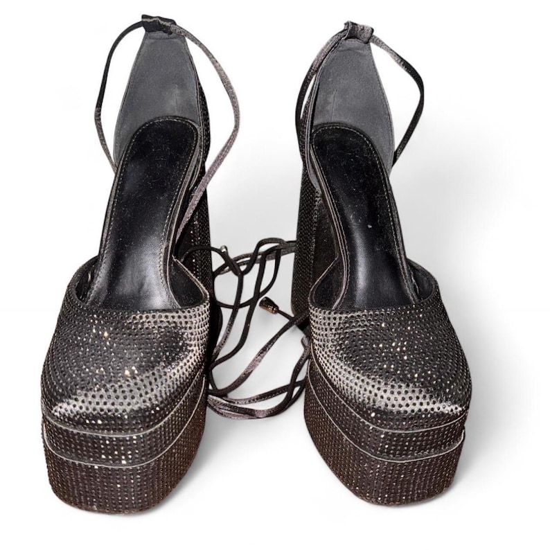 K&ouml;nnte beinhalten: Ein Paar schwarze Plateau-High Heels mit funkelnden, strass&auml;hnlichen Verzierungen. Die Schuhe haben eine dicke Plateausohle und Kn&ouml;chelriemen zum Schn&uuml;ren. Der Hintergrund ist wei&szlig;.