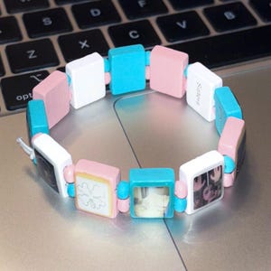Op de afbeelding: Een kleurrijke armband bestaande uit vierkante houten kralen in wit, roze en turquoise. Sommige kralen hebben afbeeldingen of ontwerpen, en andere hebben het woord "Sisters" erop gedrukt. De armband is geregen met kleine, ronde kralen.