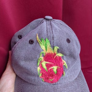 Può includere: Un cappellino da baseball grigio antracite con un disegno ricamato di un frutto del drago sul pannello frontale. Il ricamo mostra un corpo del frutto magenta vibrante con squame verde lime e gialle, accentuato da occhielli di ventilazione scuri.