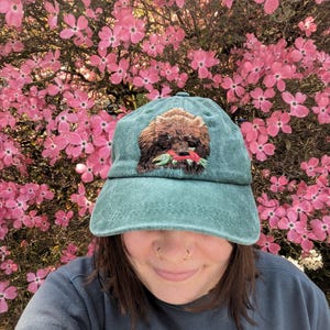 May include: A green baseball cap featuring an embroidered brown dog holding a red fish in its mouth. The cap is being worn by a person in front of a pink flowering bush.