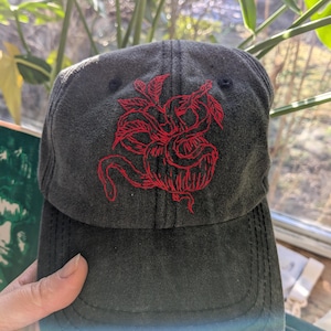 May include: A dark grey baseball cap featuring a red embroidered design of a stylized floral or botanical motif. The cap has a curved peak and a classic six-panel design, suitable for casual wear.