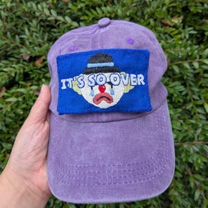 May include: A purple baseball cap with a curved peak. The cap has a blue patch embroidered with the words "IT'S SO OVER" and a crying clown face. The cap is made of a soft, textured fabric.