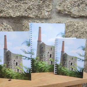 May include: Three spiral bound notebooks featuring a photograph of a stone building with a tall red brick chimney. The building is surrounded by green foliage.