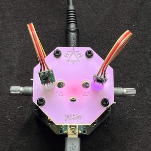 Op de afbeelding: Een close-up van een printplaat met een roze, zeshoekige toplaag. De plaat heeft twee knoppen, twee sets draden en een USB-poort. De tekst "µLSe" is zichtbaar op de plaat. De draden zijn rood, geel en bruin.