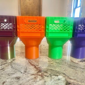 Könnte beinhalten: Vier farbenfrohe, kunststoffartige Objekte in Kistenform. Jedes hat eine andere Farbe: Burgunderrot, Orange, Grün und Lila. Die Kisten sitzen auf einem passenden, trichterförmigen Sockel. Die Objekte sind auf einer gesprenkelten Arbeitsplatte angeordnet.