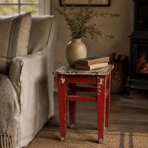 Könnte beinhalten: Ein kleiner, vierbeiniger Holzhocker mit abgenutzter roter Lackierung, unter der das weiße Holz zum Vorschein kommt. Auf der quadratischen Sitzfläche stehen eine beige Keramikvase mit grünen Zweigen und zwei gestapelte, alte Bücher.
