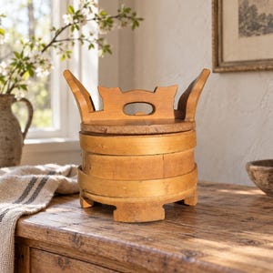 May include: A round wooden container with a lid featuring a decorative handle and two upward-curving side extensions. The container is constructed from light brown wood with visible grain and is supported by four small, integrated wooden feet.
