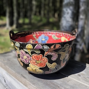 May include: A decorative black bowl with colorful floral designs. The bowl has a red interior with a floral pattern and two small handles. The exterior features flowers in shades of orange, purple, yellow, and blue.