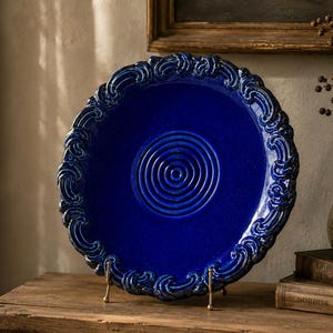 May include: A deep cobalt blue ceramic plate featuring a raised, swirling scroll pattern along the outer rim and a series of concentric circular ridges embossed in the centre. The plate is displayed upright on a thin, gold-toned metal stand.