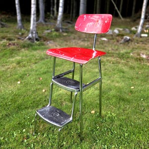 May include: A vintage red step stool with a small backrest. The stool has a chrome frame and two black, textured steps. The stool is set on a grassy lawn with trees in the background.