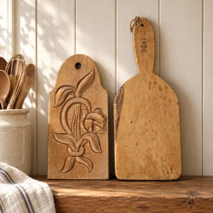 May include: Two light brown wooden butter moulds. The left mould features a carved floral design and a circular hole at the top. The right mould has a long handle with a loop of twine and the stamped text 1890. Both items have a weathered finish.