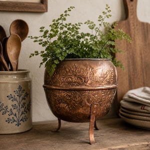 Peut inclure: Un pot de fleurs en cuivre à trois pieds avec des motifs floraux en relief, rempli de plantes vertes. Le pot est posé sur une étagère en bois, avec un pot en céramique et des cuillères en bois en arrière-plan.