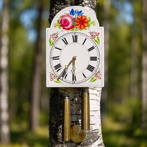 Könnte beinhalten: Eine weiße Wanduhr mit floralem Design und römischen Ziffern. Die Uhr hat goldfarbene Gewichte und ein Pendel. Die Uhr ist an einem Baumstamm in einer Waldlandschaft befestigt.