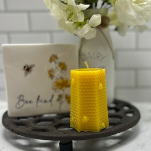May include: A yellow beeswax candle with a honeycomb pattern and a small bee design. The candle is sitting on a black metal trivet with a white ceramic tile that says "Bee kind" and a floral design.